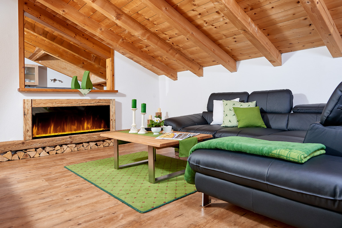 Alpglück Wohnzimmer mit Couch, Couchtisch und Kamin sowie grünen Akzenten durch Kerzen, Decke und Teppich